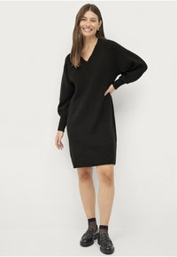 Zwarte gebreide jurk met een V-hals, losse mouwen en geribbelde zoom. Gecombineerd met zwarte loafers en maillot sokken. Glad textuur.