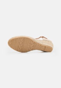 Sandalia de cuña con parte superior trenzada en color beige, que presenta una suela de goma texturizada y una correa en el tobillo marrón con detalles de hardware.