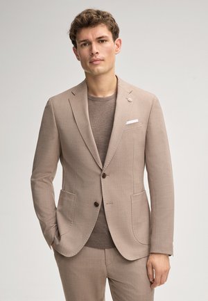 Jonge man die een beige pak draagt met een bruine trui eronder, staand met één hand in zijn zak tegen een eenvoudige lichte achtergrond.