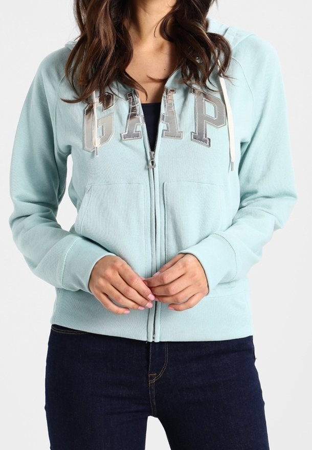 Sweat à capuche zippé bleu clair avec le logo "GAP" en lettres argentées et capuche avec cordon de serrage. Comprend des poches avant et des poignets côtelés, fabriqué en tissu doux.