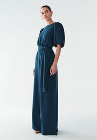 Turkooise, vloerlang jurkje met korte pofmouwtjes, een strak korset en een ceintuur die in een strik wordt vastgemaakt. Gladde, vloeiende stof zonder zichtbare patronen.