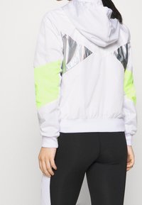 Chaqueta blanca con capucha, acentos en verde neón y paneles plateados reflectantes. Presenta puños y cintura elásticos, combinada con leggings negros.