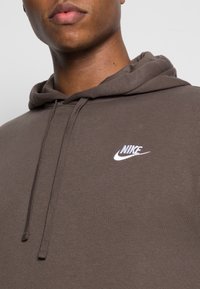Brązowa bawełniana bluza z kapturem z przednią kieszenią typu kangur, regulowanym kapturem ze sznurkiem oraz białym haftem logo Nike po lewej stronie klatki piersiowej.