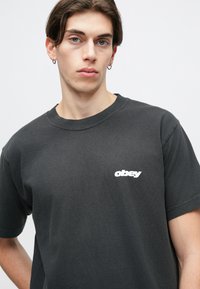 T-shirt de algodão preto com gola redonda e mangas curtas, apresentando um logótipo branco "obey" no peito. Textura suave.