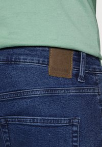  Jeans de denim azul com um patch de couro castanho exibindo "ONLY & SONS." O tecido possui uma leve textura, e as costuras estão visíveis ao longo das emendas.