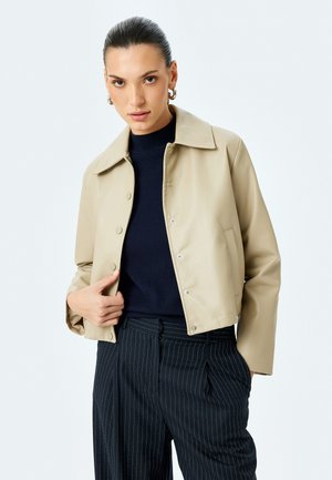 Chaqueta corta beige hecha de un material suave, con un gran cuello, cierre de botones y bolsillos laterales, combinada con un suéter oscuro y pantalones a rayas.
