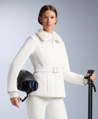 Jachetă albă de schi cu guler de blană, textură ribbată și detaliu cu curea. O cască neagră cu vizor colorat și un schi pole în mână.