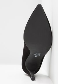 Zapato de tacón alto de ante negro con punta afilada, tacón delgado y suela negra lisa, que presenta una etiqueta que dice "Cuero en la parte superior, Hecho en España."