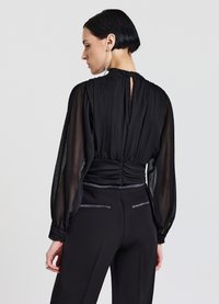 Blusa nera trasparente con maniche lunghe e voluminose, schiena arricciata e scollatura rotonda, abbinata a pantaloni neri sartoriali con accenti discreti.