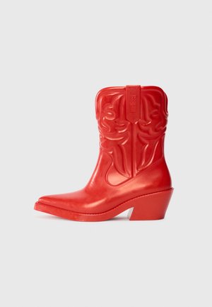 Botte de cowboy rouge avec un talon haut et évasé. Fabriquée en matériau brillant avec des motifs en relief de style western sur la tige. Forme de bout pointu.