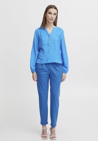 Blouse bleu à manches longues avec un tissu texturé et un décolleté en V, assortie à un pantalon bleu. Le modèle porte des sandales blanches à lanières.