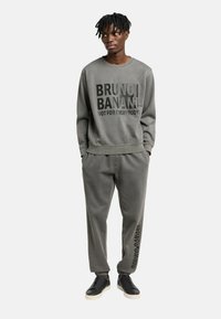 Grijze sweatshirt met vetgedrukte zwarte tekst, gecombineerd met bijpassende grijze joggingbroeken. Soepele stof en een ontspannen pasvorm. Zwarte instapschoenen maken de look compleet.