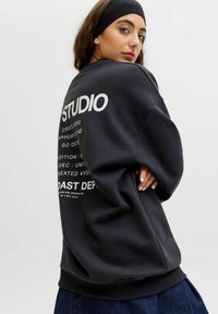 Černá mikina s kulatým výstřihem, volným střihem a bílým grafickým textem na zadní straně. Hladký materiál s ribovanými manžetami a lemem.