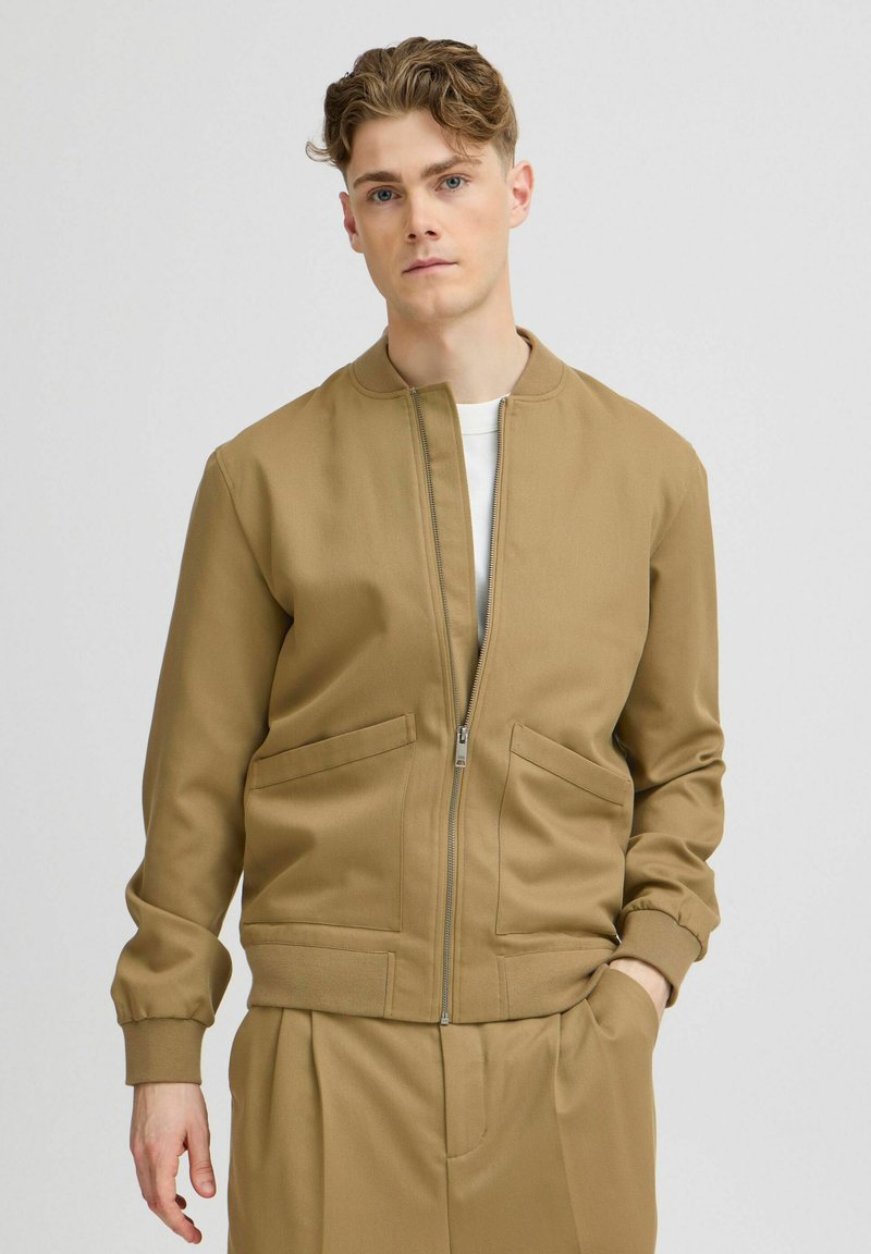 Khaki bomberjack met een rits aan de voorkant, geribbelde manchetten en twee zijzakken. Draagt over een wit shirt, gladde textuur.