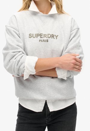Lysegrå Superdry Paris sweatshirt båret over en hvid skjorte med krave og opkrogede ærmer, kombineret med sorte bukser.