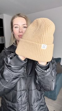 Barna kötött sapka, bordás textúrával, egy kis címkével, amelyen egy logó található. Egy fekete kabátot viselő személy előtt tartják.