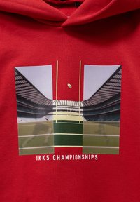 Sudadera roja con capucha que presenta un gráfico de un estadio deportivo con un campo verde, postes de gol y una pelota de rugby, etiquetado como "IKKS CHAMPIONSHIPS".