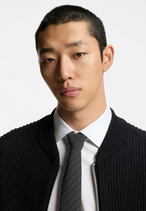 Giacca bomber nera a coste con cerniera argento, indossata sopra una camicia bianca con motivo e una cravatta grigia a righe. Primo piano, sfondo neutro.