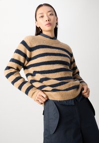 Pulover bej din lână cu dungi orizontale bleumarin, guler rotund și manșete dublate, purtat împreună cu pantaloni negri care au buzunare laterale.