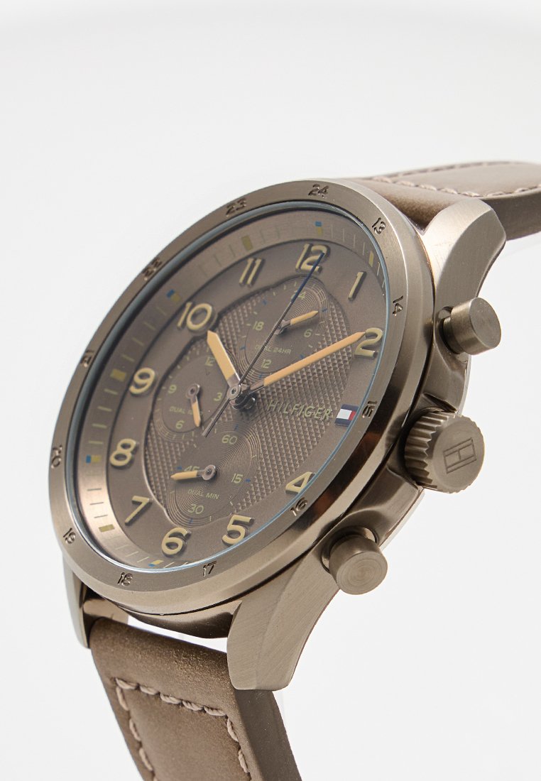 Brązowy zegarek chronograficzny z teksturowaną tarczą, dużymi numerycznymi znacznikami godzinowymi i metalową kopertą. Posiada beżowy skórzany pasek oraz dwa przyciski.
