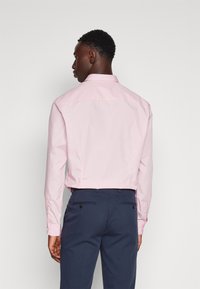 Chemise habillée rose clair avec un col et des manches longues, associée à un pantalon sombre avec des détails de boutons et une texture de tissu lisse.