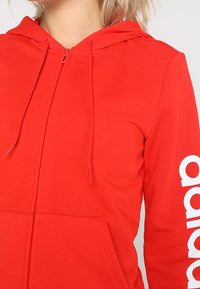 Felpa rossa con zip realizzata in tessuto morbido, dotata di cappuccio, cordini e testo del logo bianco lungo la manica sinistra.