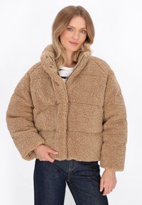 Tan faux fur jakke med høj krave og trykknapper, der har et firkantet design og tekstureret overflade, båret over en stribet skjorte.