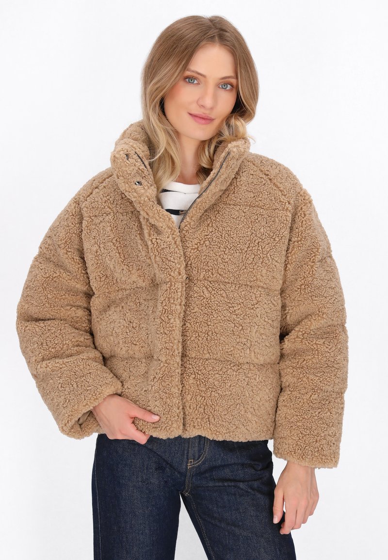 Tan faux fur jakke med høj krave og trykknapper, der har et firkantet design og tekstureret overflade, båret over en stribet skjorte.