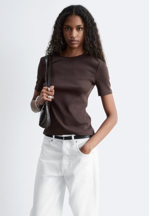 Joven mujer con cabello rizado y largo lleva una blusa marrón de manga corta, pantalones blancos, cinturón negro, pendientes de aro, anillos, pulseras y sostiene un bolso negro de hombro.