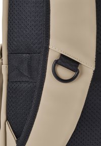 Beige hátizsák anyag fekete hálós részletekkel, gyűrűs rögzítéssel és megerősített varratokkal a tartósság érdekében.