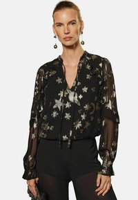 Blusa nera trasparente con stampa di stelle e lune argento, dettagli a volantino, scollatura a V e maniche lunghe; abbinata a pantaloni neri in tessuto.