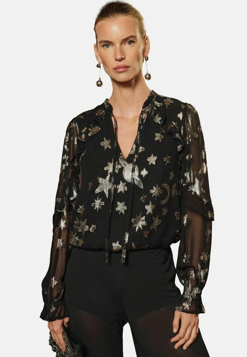 Blusa nera trasparente con stampa di stelle e lune argento, dettagli a volantino, scollatura a V e maniche lunghe; abbinata a pantaloni neri in tessuto.