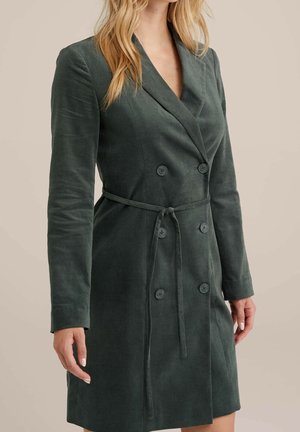 Kvinde iført en mørkegrøn, dobbeltradet, bæltebelagt velourblazer-kjole med lange ærmer og reverskrave.