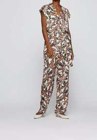 Mono floral en crema con patrones de hojas en naranja, rosa y negro. Presenta mangas cortas, cintura ceñida con cinturón y pantalones de pierna ancha.