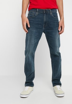 Donkerblauwe denim jeans met een rechte pasvorm, vijf zakken en een rits. G gedragen met witte sneakers en een rood shirt voor contrast.