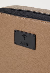 Skórzana torba o teksturowanej, beżowej powierzchni z czarnym zamkiem błyskawicznym. Posiada czarną metalową tabliczkę z logo "JOOP!" oraz dekoracyjny emblemat.
