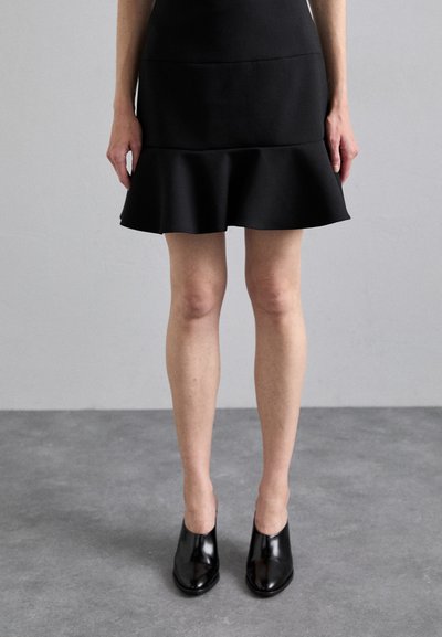 Jupe noire ajustée avec un ourlet évasé, en tissu lisse. Assortie à des mules à talons brillants noires. Fond neutre.