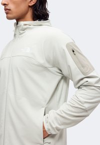 Personne portant une veste North Face gris clair avec une poche zippée sur le haut du bras et les mains dans les poches latérales.