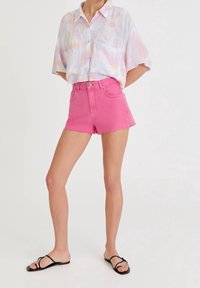 Shorts en jean roses courts avec ourlet effiloché, associés à une chemise ample à boutons multicolore pastel. Portés avec des sandales plates noires.