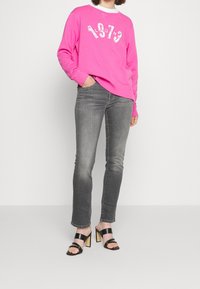 Ljusrosa sweatshirt med grafik "1973", kombinerad med grå tighta jeans och svarta högklackade sandaler med remmar. Enkelt design, avslappnad passform.