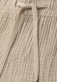 Pantalon en tissu beige texturé avec une taille élastique, une poche avant et un cordon plat noué en un simple nœud.