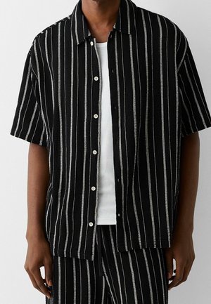 Camisa de rayas en blanco y negro de manga corta con cuello, que presenta una tela texturizada y cierres de botón. Llevada sobre una camiseta sin mangas blanca.