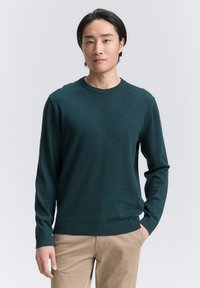 Suéter de cuello redondo en verde oscuro de punto ligero, con mangas largas y puños acanalados, combinado con pantalones beige.