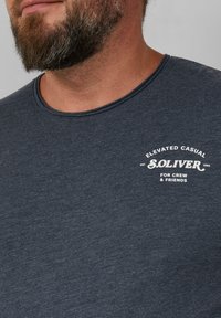 Dunkelgraues Baumwoll-T-Shirt mit rundem Halsausschnitt, bedruckt mit weißem Text: "ELEVATED CASUAL S.OLIVER FÜR CREW & FRIENDS."