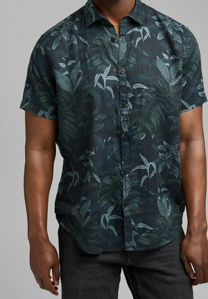 Uomo con una camicia scura a maniche corte con bottoni e motivo di foglie tropicali blu e verdi e pantaloni scuri.