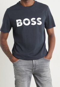 Mörk marinblå t-shirt i bomull med fet vit text "BOSS". Korta ärmar och rund halsringning. Bärs med ljusgrå jeans.