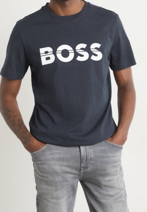 Camiseta de algodón en azul marino oscuro, con letras en blanco en negrita que dicen "BOSS". Con mangas cortas y cuello redondo. Se lleva con jeans de color gris claro.