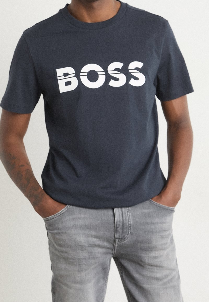 Mörk marinblå t-shirt i bomull med fet vit text "BOSS". Korta ärmar och rund halsringning. Bärs med ljusgrå jeans.