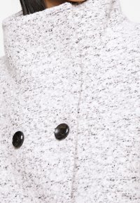 Detail bílo-šedého texturovaného kabátu s vysokým límcem a dvěma černými knoflíky vpředu.