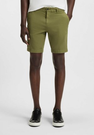 Olivgrüne Shorts mit geradem Schnitt, Taillenbund mit Knopfverschluss und umgeschlagenem Saum. Getragen mit schwarzen Sneakers vor einem einfarbigen Hintergrund.
